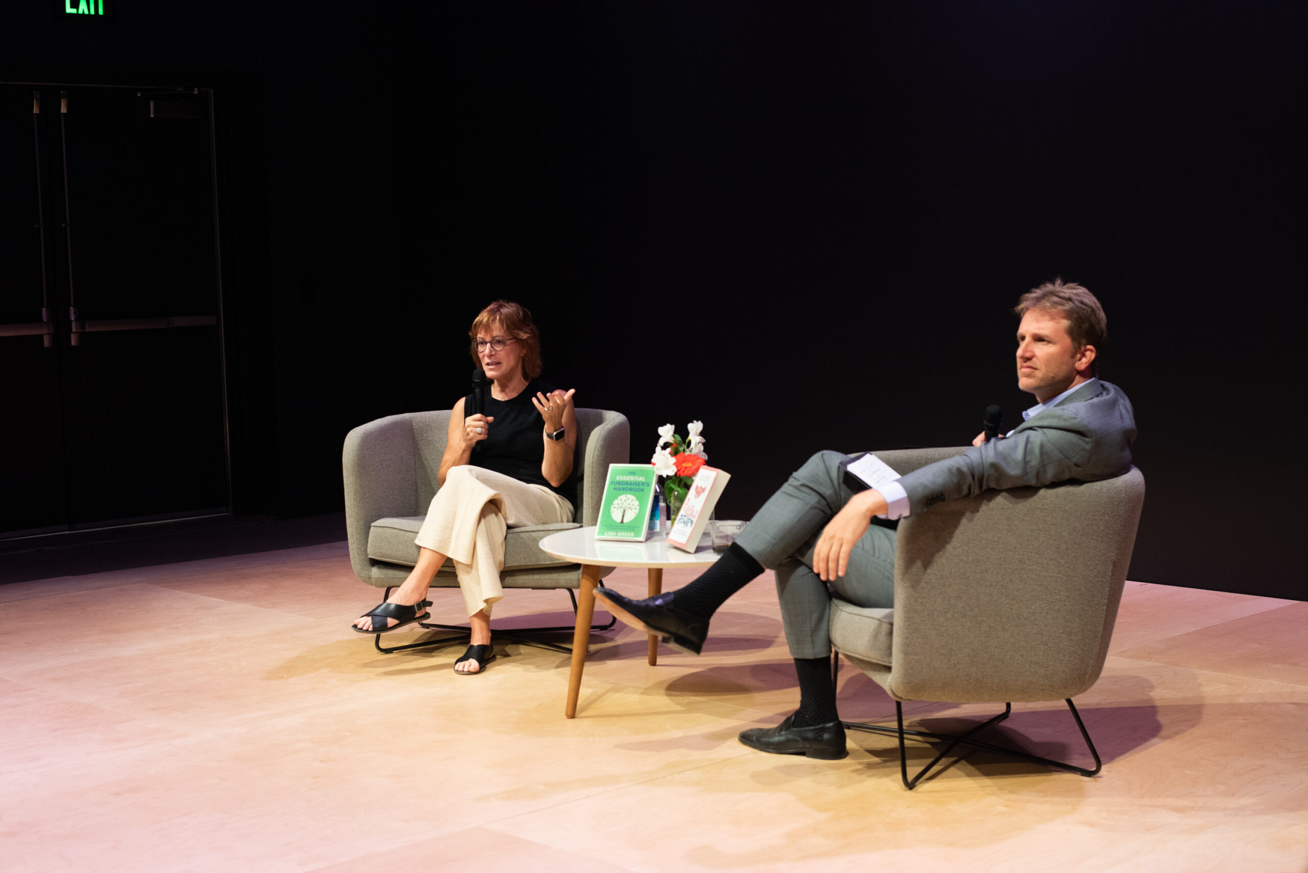 Author speaking on stage at a book launch event, seated in a grey armchair next to a table displaying her books.